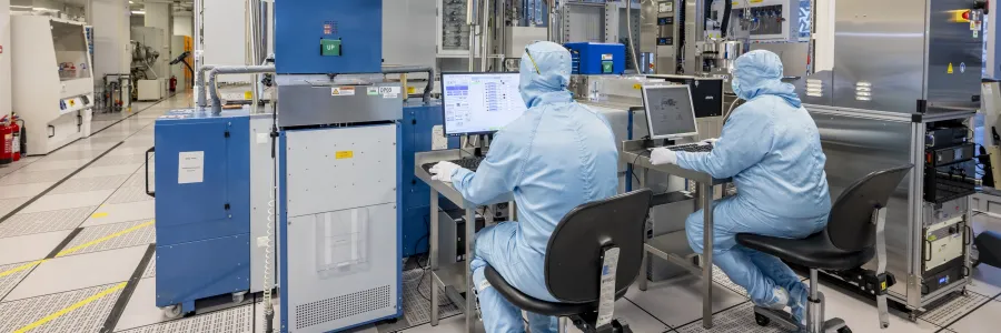 Two men in full cleanrooms uniforms are sitting at the desks working with equipment