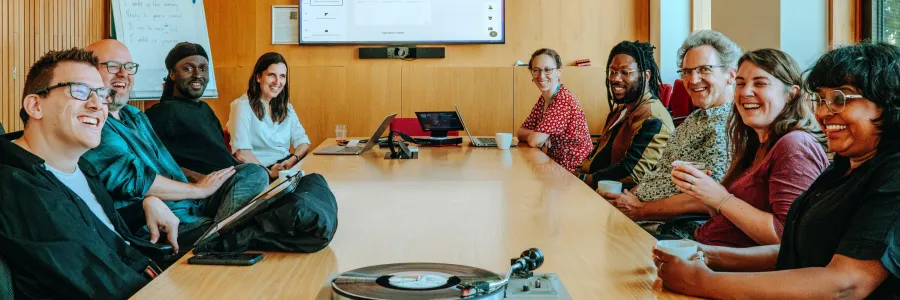  A group of nine people is seated around a wooden conference table, smiling and engaged in a meeting. Laptops are open, and a turntable with a vinyl record sits at the front of the table. A large screen behind them shows a web interface, and a whiteboard with notes is visible. The mood is friendly and collaborative in a modern, well-lit room.