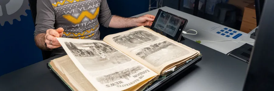 A man digitizes a large illustrated book using a professional overhead camera setup and bright studio lights. The workspace includes advanced imaging equipment, highlighting attention to detail and preservation.