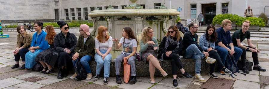  A group of people is sitting on the edge of a circular fountain in an outdoor plaza. They are engaged in casual conversations, with the Southampton Art Gallery and banners visible in the background.