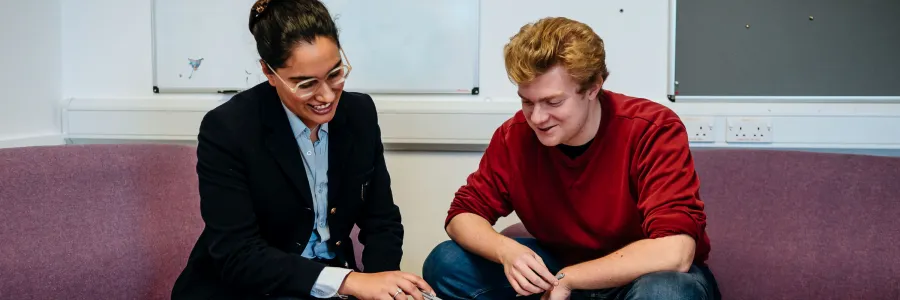 Two people are sitting on a couch in an office space, reviewing a document or booklet together. The woman is smiling and pointing at the material, while the man attentively listens and looks on.