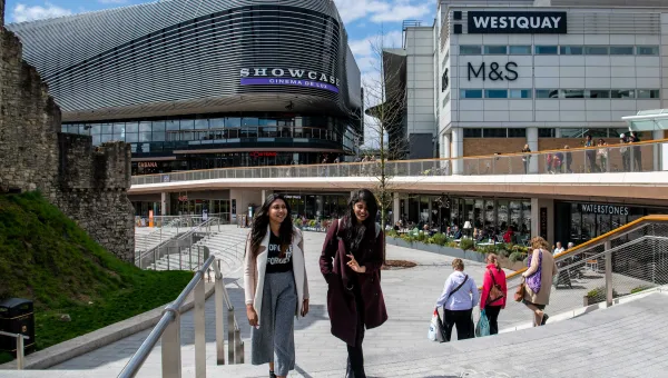 Two students talk whilst walking across a large paved area with several large shops.