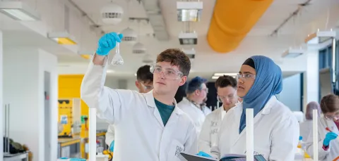 Two students wearing lab coats and protective glasses conduct an experiment. One holds up a glass beaker, while the other takes notes. They are in a laboratory, surrounded by a variety of chemistry equipment.