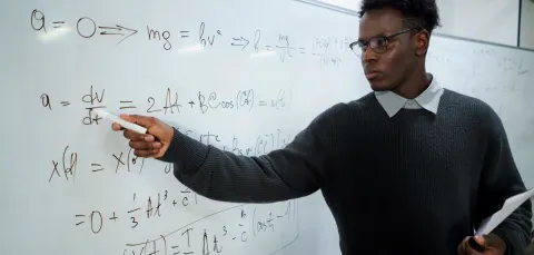 A maths teacher writes on a white board and looks intently at the equations. We imagine he is in front of a classroom of pupils.