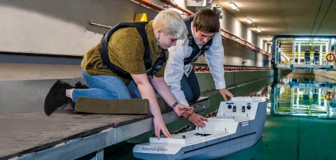 A student crouches down to place a model boat into the water of the university towing tank, a researcher at her side