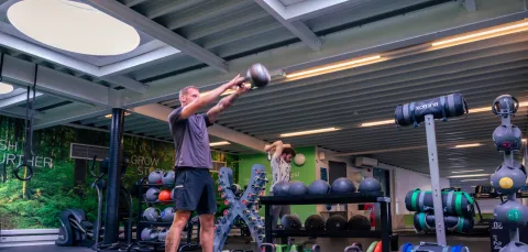 A man lifting a dumbell straightout in front of his body. There are lots of weights and equipment behind.