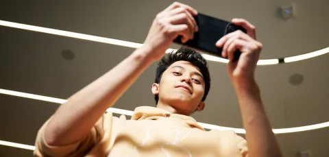 A student is looking down at the camera while using their smartphone, which they are holding horizontally. The student is wearing a pale yellow hoodie, and stood in a corridor which is well-lit by several fluorescent lighting strips.