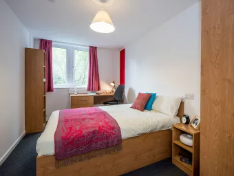 Bedroom with a neatly made double bed and nightstand. A wooden desk and open window are visible in the background.