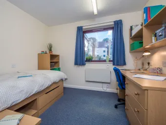 A window lets light into a bedroom where a neatly made bed and desk can be seen.