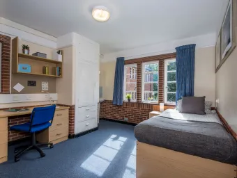 Light from a large window fills a spacious bedroom with desk, chair and neatly made bed.