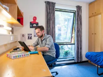 A student sits looking at a tablet computer while working at a desk. A bed and wardrobe can be seen behind him, an open window lets light into the room.