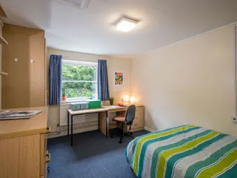 A large student bedroom. The room contains a double bed with striped covers, a large desk, a wooden wardrobe and chest of drawers.