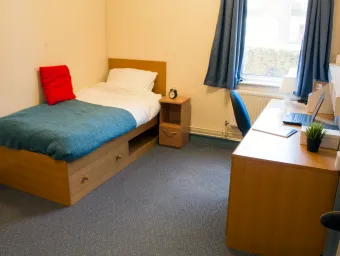 A spacious bedroom. The floor is blue carpeted and a neatly made single bed is visible against one wall. On the other side of the room is a wooden desk with office chair and a large window.