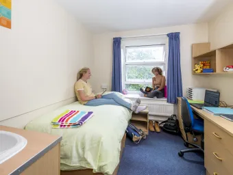 Two students chatting in a bedroom. One sits browsing with her tablet on a single bed while the other looks up from their book, sat on the windowsill.