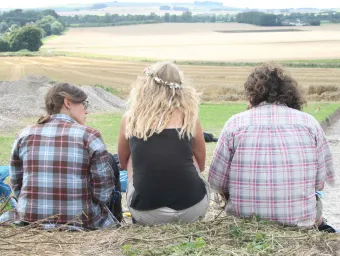 Three people sit on a hill overlooking a large open area of countryside at Avebury, a site of ancient settlement.
