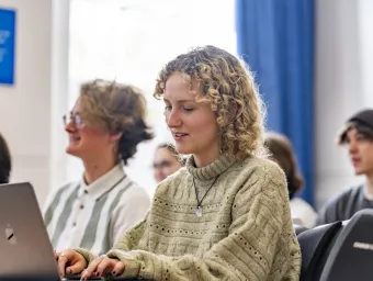 A student is sitting at a desk, typing on their laptop. They have blonde curly hair and a beige knitted jumper. Other students are sitting in the same room but are out of focus. They all face the same direction and are smiling.