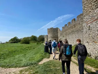 A group of history students walk beside the outside wall of the Hampshire medieval fortress Portchester Castle, as part of a field trip. The 2 students closest to us are in discussion.