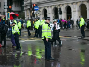 A number of police officers monitor crowds of people on a rainy London street. They are wearing high visibility yellow jackets.