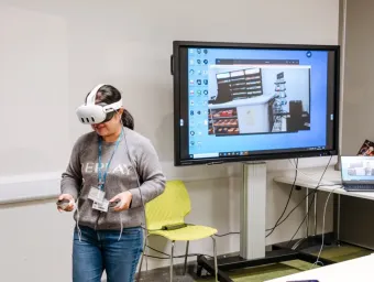 Community hub event participant demonstrates virtual reality headset. A large monitor and laptop are visible in the background.