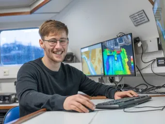 Daniel Hampton is sitting at the work station on Research Vessel Callista. There are screens displaying coulourful charts in the background.