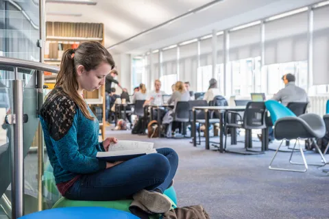 A student sits cross-legged reading a book in a bright, airy library. Behind her are shelves of books and other students working at desks.