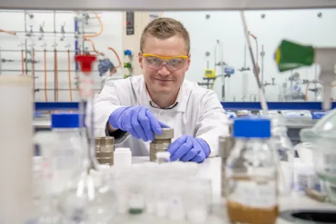 A chemistry researcher in goggles, gloves and white coat works with equipment in the lab