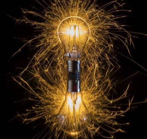 An illuminated light bulb with a vibrant shower of golden sparks radiating around it, resting on a reflective surface with a visible reflection below.