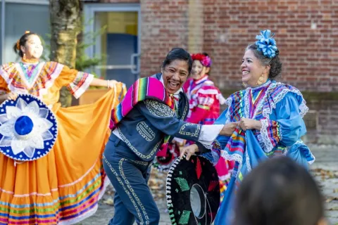 People are celebrating outside. They wear traditional Mexican dress and are smiling happily. In the foreground, one dancer is wearing a blue suit with a yellow decorative print following the seams, and a colourful striped garment over a shoulder. They are laughing and holding the hand of another dancer who is wearing a blue dress with a matching hair bow. The dress has lace trims and rainbow decoration at the collar and cuffs. They are holding a black wide-brimmed hat decorated with green and red. 