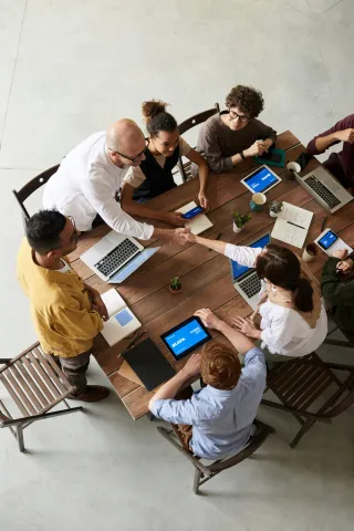 A top-down view of a diverse group of six professionals seated around a wooden table with laptops and tablets, engaged in discussion during a collaborative meeting in a modern workspace.