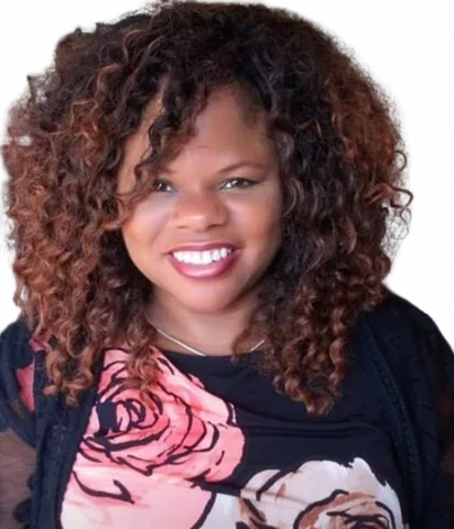 A photo of public contributor, Traci Carroll. She is wearing a black top with oversized pink and white roses. She has beautiful curly natural hair.