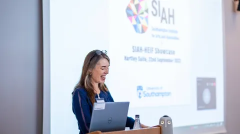 A person is speaking at a lectern with an open laptop in front of them. There is a projector screen behind them which is displaying the SIAH logo. 