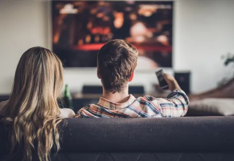 A couple sitting on a sofa, watching a wall mounted TV. The man is pointing the remote at the screen.