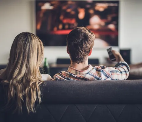 2 people sitting on a soda watching a large wall mounted TV. The man is pointing the remote at the screen.