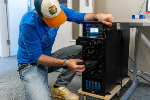 A person is inserting or removing a hard drive from a multi-bay server or data storage system in a tech workspace. They are wearing a blue shirt, jeans, and a cap with the label "Outdoor."