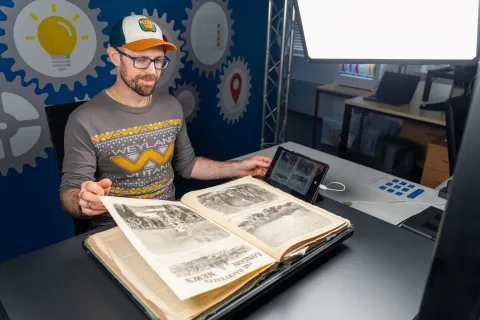 A man digitizes a large illustrated book using a professional overhead camera setup and bright studio lights. The workspace includes advanced imaging equipment, highlighting attention to detail and preservation.