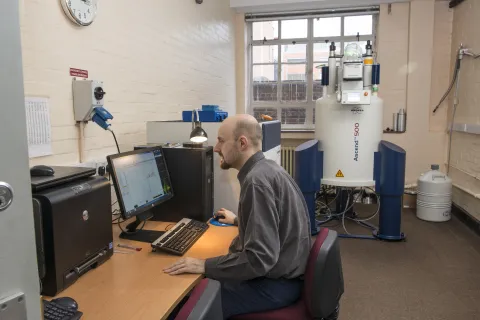 A man working  on a computer in a room with a Nuclear Magnetic Resonance machine in the background