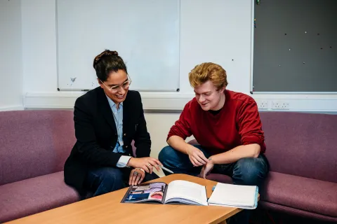 Two people are sitting on a couch in an office space, reviewing a document or booklet together. The woman is smiling and pointing at the material, while the man attentively listens and looks on.