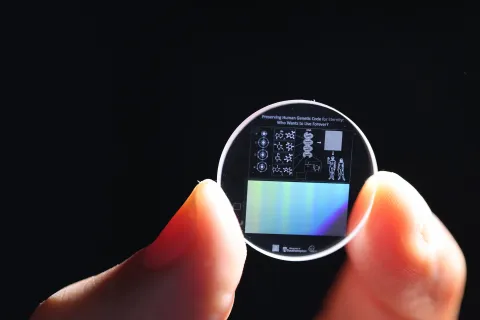 Zoomed in view of a hand holding a 5D memory crystal between two fingers. The memory crystal is a small clear disc with diagrams of human DNA on the surface.