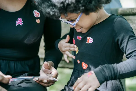 A young child is choosing stickers from a sheet held by an adult. The child is adding the colourful stickers to their top.  