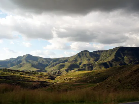 A landscape of rolling hills with long grass in the Lesotho Highlands. There is a mountain range on the horizon, and the sky is mainly cloudy with some clear blue areas.