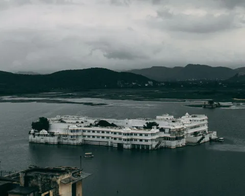 The Taj Lake Palace in Udaipur, Rajasthan, India. It is a rainy day and there are dark clouds surrounding the hills in the distance.