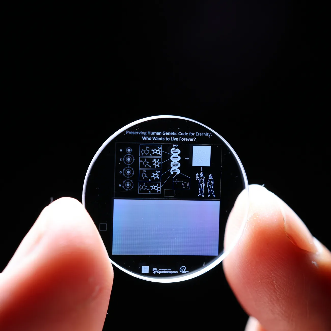 Zoomed in view of a hand holding a 5D memory crystal between two fingers. The memory crystal is a small clear disc with diagrams of human DNA on the surface. 