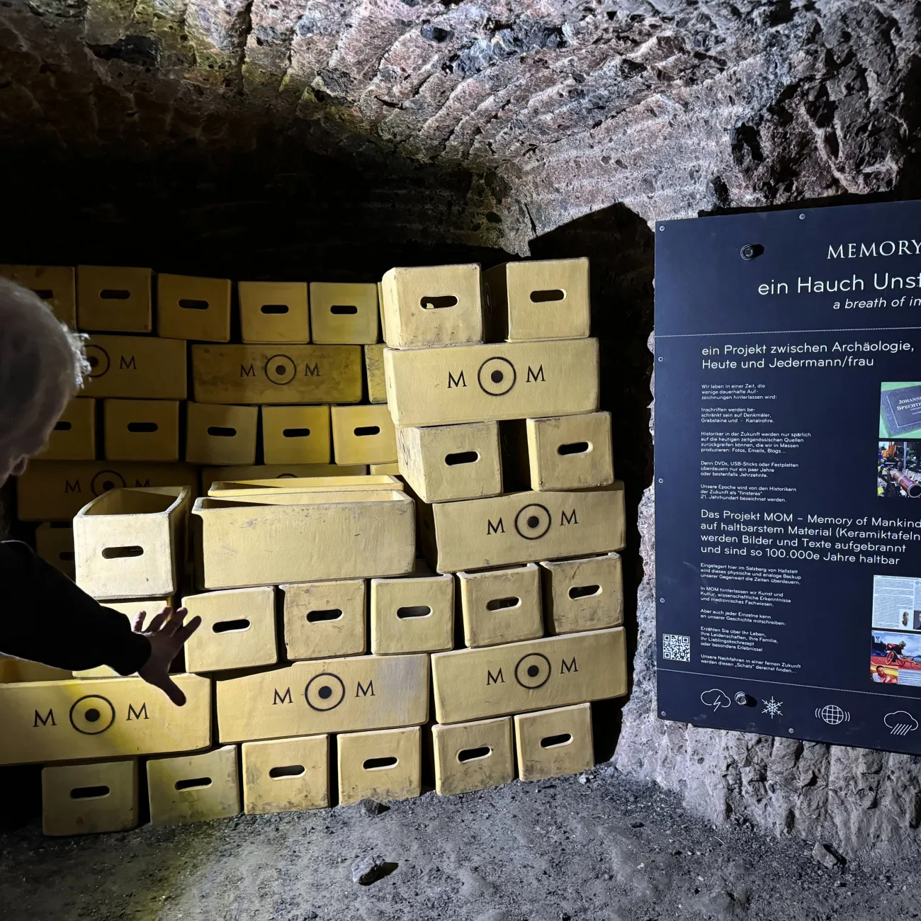 A salt cave in the Memory of Mankind archive. There are yellow time capsule boxes stacked against the brick walls and a large blue sign with visitor information written in German.