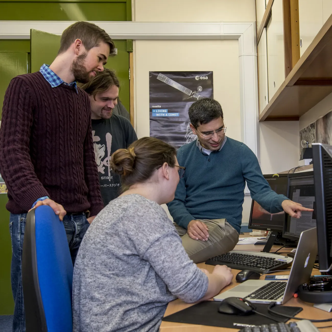 A group of 4 people sat at and stood around a desk in a laboratory with several computers, laptops and monitors. One of the people is sat on the desk and pointing at one of the monitors, while the other three people are looking at what they are pointing at.