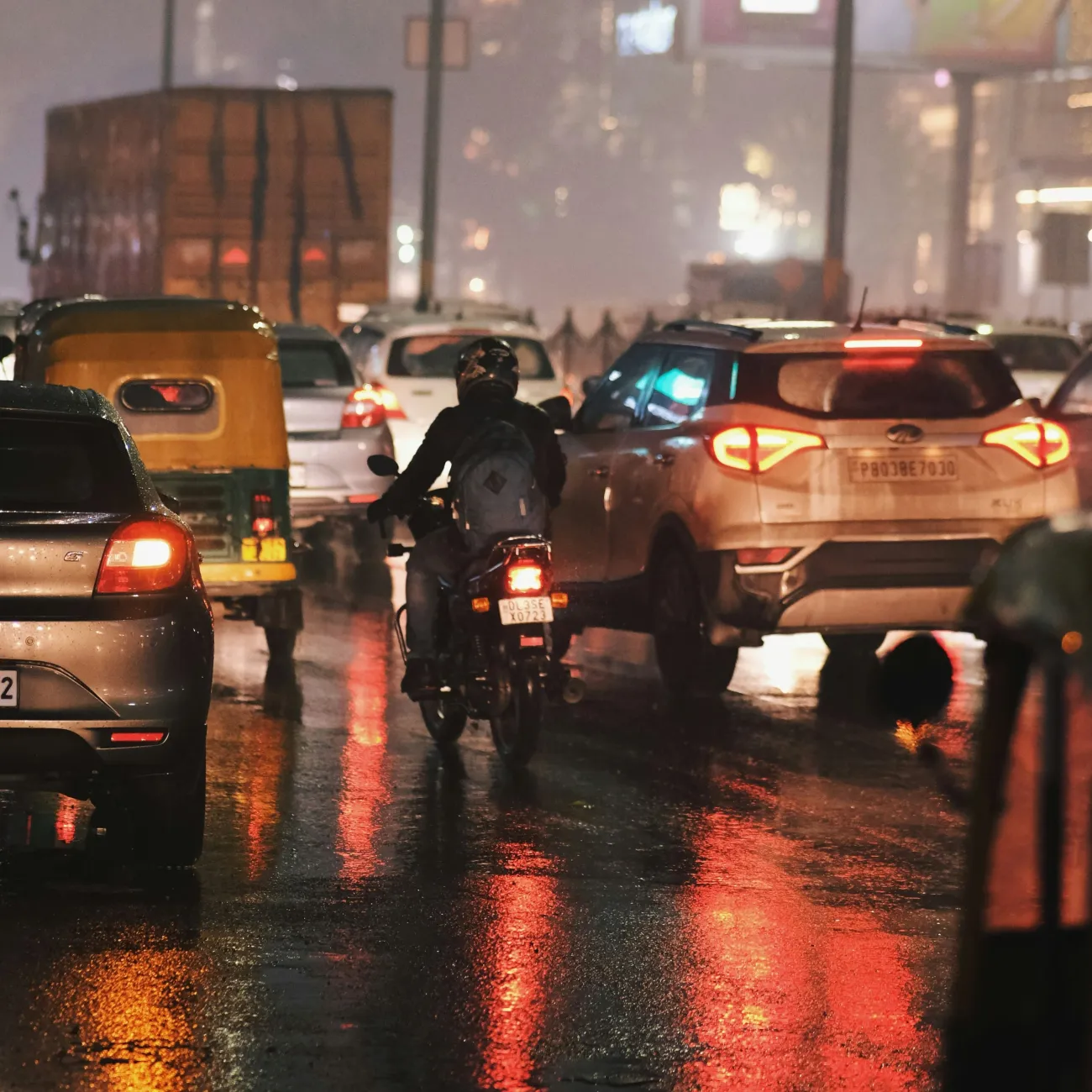 A busy road in South Delhi, India at night. It is raining and there are multiple cars and motorbikes on the road.