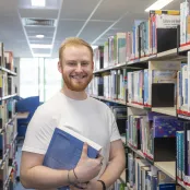 Public Health student, Jack standing between library shelves holding a book. He is smiling at the camera.