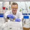 A chemistry researcher in goggles, gloves and white coat works with equipment in the lab