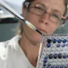 A close-up of a scientist in a white lab coat pipetting dark blue liquid into a 96 well lab plate