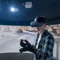 A student wearing a virtual reality headset and gloves, standing in front of a 360 degree video wall displaying what he can see.