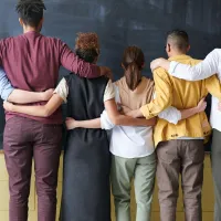 A diverse group of nine people stand in a row with their arms around each other's shoulders, facing a blackboard in a classroom, dressed in an array of casual to smart-casual attire.